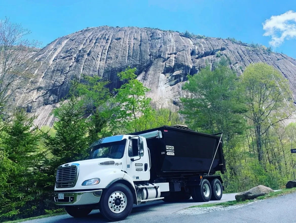Dump truck parked beside a large rocky cliff and green trees.